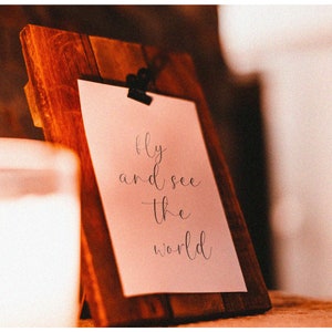 K&ouml;nnte beinhalten: Ein Holzbildrahmen mit einer wei&szlig;en Karte darin. Die Karte tr&auml;gt den Text "Fly and see the world" in schwarzer Kursivschrift.