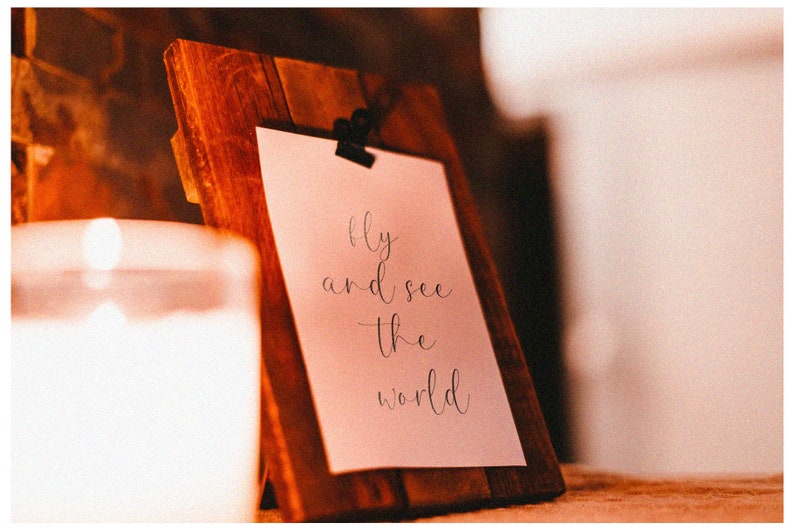 K&ouml;nnte beinhalten: Ein Holzbildrahmen mit einer wei&szlig;en Karte darin. Die Karte tr&auml;gt den Text "Fly and see the world" in schwarzer Kursivschrift.