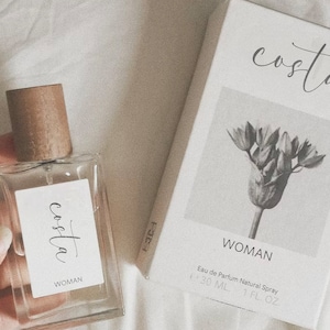 May include: A clear glass bottle of perfume with a brown wooden cap. The bottle has a white label with the word "costa" written in cursive and the word "WOMAN" underneath. The bottle is sitting on a white surface. The bottle is 30 ml or 1 fl oz.