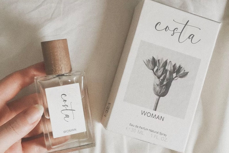 May include: A clear glass bottle of perfume with a brown wooden cap. The bottle has a white label with the word "costa" written in cursive and the word "WOMAN" underneath. The bottle is sitting on a white surface. The bottle is 30 ml or 1 fl oz.