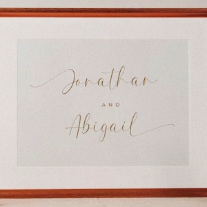 K&ouml;nnte beinhalten: Ein gerahmter Druck mit den Namen "Jonathan" und "Abigail", die in Schreibschrift in goldenen Buchstaben auf wei&szlig;em Hintergrund geschrieben sind.
