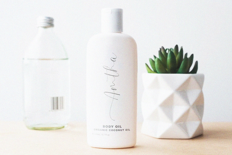 K&ouml;nnte beinhalten: Wei&szlig;e Flasche Amitha Bio-Kokosnuss-K&ouml;rper&ouml;l mit schwarzem Etikett und dem Text "BODY OIL ORGANIC COCONUT OIL 100 ml / 3.37 fl oz".