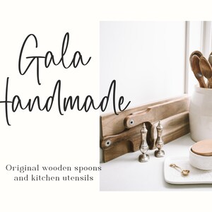 May include: A white ceramic jar filled with wooden spoons and kitchen utensils. The jar is sitting on a white countertop next to a wooden cutting board and two silver salt and pepper shakers. The text "Gala Handmade" is written in black cursive script on a white background. The text "Original wooden spoons and kitchen utensils" is written in black text below the cursive script.