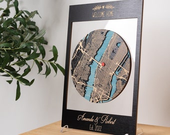 May include: A black wooden frame with a circular map inside. The map is of a city with blue water and brown land. A red heart pin is placed on the map. The frame has the text "Welcome Home" and "Amanda & Robert Est. 2022" on it.