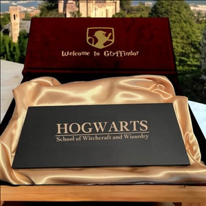 May include: A black rectangular plaque with the words "HOGWARTS School of Witchcraft and Wizardry" in gold lettering, resting on a gold satin cloth inside a burgundy box. The box lid reads "Welcome to Gryffindor".
