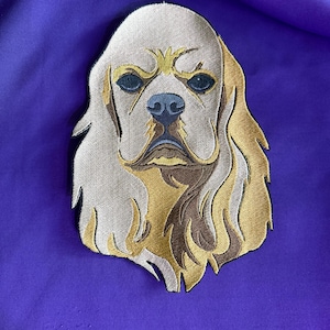May include: Embroidered patch featuring a detailed portrait of a Cavalier King Charles Spaniel dog. The patch showcases a cream-colored face with brown and gold accents, dark blue eyes, and a black nose. The patch is set against a purple background.