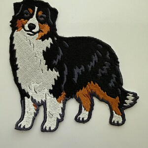 May include: Embroidered patch featuring a standing Australian Shepherd dog. The patch showcases the dog's distinctive markings: black, white, and brown fur. The dog's features are detailed with thread, creating a textured appearance. Ideal for jackets, bags, or other fabric items.
