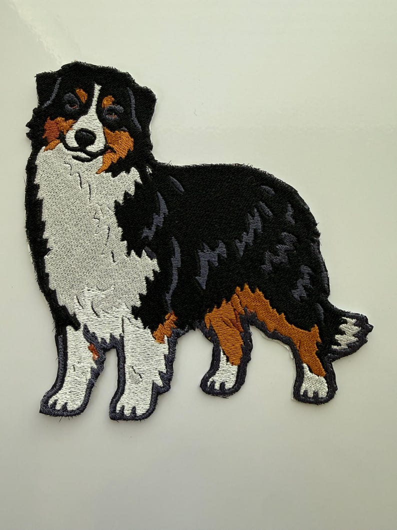 May include: Embroidered patch featuring a standing Australian Shepherd dog. The patch showcases the dog's distinctive markings: black, white, and brown fur. The dog's features are detailed with thread, creating a textured appearance. Ideal for jackets, bags, or other fabric items.
