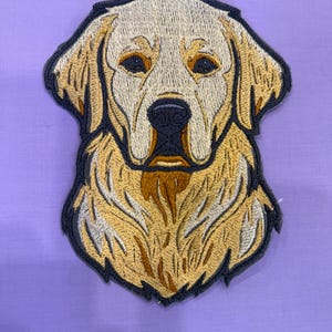 May include: Embroidered patch depicting the head of a Golden Retriever dog. The patch has a cream-coloured face, with brown and black accents. The dog's fur is a mix of gold and cream threads, outlined with a dark blue border. The patch is on a lavender background.