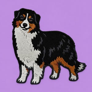 May include: Embroidered patch featuring a standing Australian Shepherd dog. The patch showcases the dog's distinctive markings: black, white, and brown fur. The dog is set against a solid purple background, highlighting the detailed stitching and design.