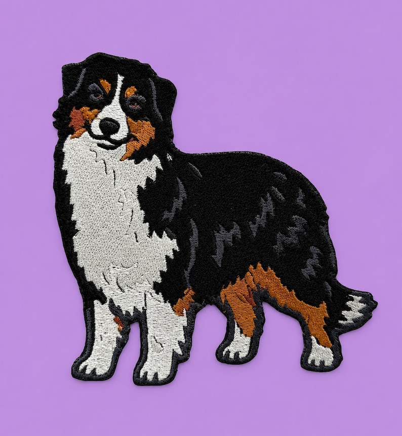 May include: Embroidered patch featuring a standing Australian Shepherd dog. The patch showcases the dog's distinctive markings: black, white, and brown fur. The dog is set against a solid purple background, highlighting the detailed stitching and design.