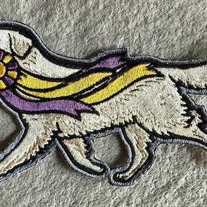 May include: Embroidered patch of a Golden Retriever dog in a walking pose. The dog is cream-colored with a black outline. It wears a yellow and purple ribbon and a rosette. The patch is approximately 4 inches long.