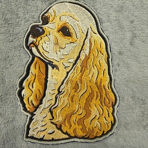 May include: Embroidered patch of a golden Cocker Spaniel dog in profile. The patch has a detailed design with shades of gold, brown, and black, outlined with a black border. The dog's fur is textured, and the patch is set against a grey background.