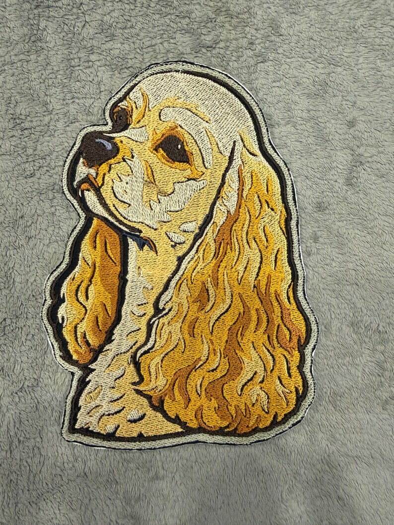 May include: Embroidered patch of a golden Cocker Spaniel dog in profile. The patch has a detailed design with shades of gold, brown, and black, outlined with a black border. The dog's fur is textured, and the patch is set against a grey background.