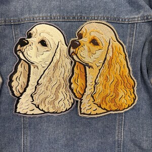 May include: Two embroidered Cocker Spaniel dog patches in cream and gold, with black detailing, are attached to a denim jacket. The patches depict the dogs' heads, highlighting their long ears and expressive eyes.