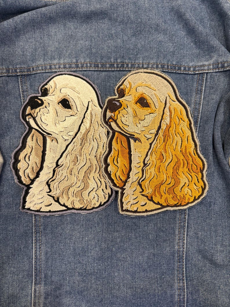 May include: Two embroidered Cocker Spaniel dog patches in cream and gold, with black detailing, are attached to a denim jacket. The patches depict the dogs' heads, highlighting their long ears and expressive eyes.