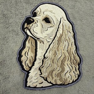 May include: Embroidered patch depicting a Cocker Spaniel dog. The patch shows the dog's head and shoulders, with cream and tan hues, and a black outline. The dog's fur is intricately stitched, giving it a textured appearance. The patch is likely for clothing or accessories.