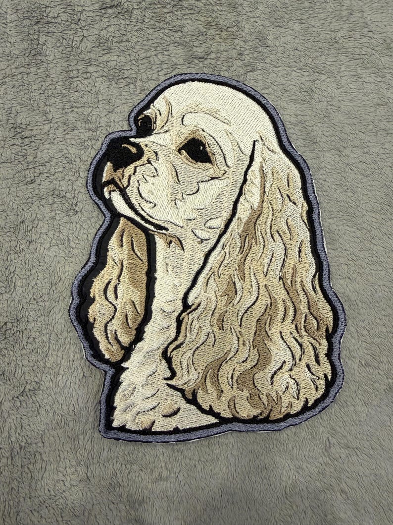 May include: Embroidered patch depicting a Cocker Spaniel dog. The patch shows the dog's head and shoulders, with cream and tan hues, and a black outline. The dog's fur is intricately stitched, giving it a textured appearance. The patch is likely for clothing or accessories.