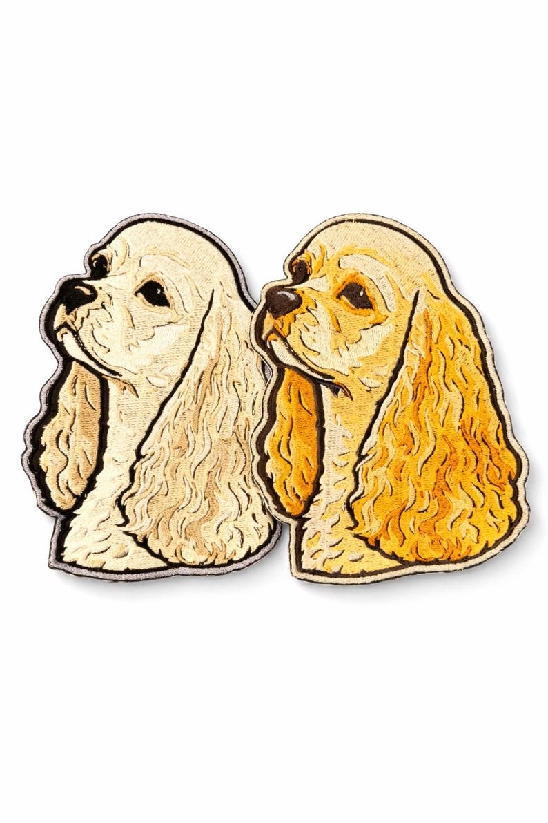 May include: Two embroidered Cocker Spaniel dog head patches. One is cream-colored, the other golden yellow. Both have black outlines and detailed facial features. The patches are designed to be sewn or ironed onto fabric.