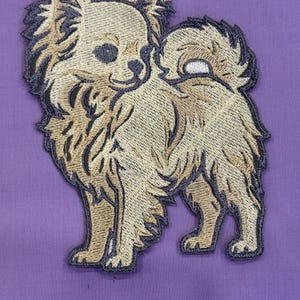 Puede incluir: Parche bordado de un perro chihuahua marrón y negro. El perro está de pie con la cola enroscada. El parche tiene un borde negro y está sobre un fondo morado. El pelaje del perro está detallado con varios tonos de marrón.