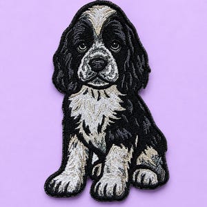 May include: An embroidered patch featuring a sitting puppy with black and white fur. The dog has long, floppy ears, a white blaze on its forehead, and a white chest. The embroidery uses detailed stitching to define the facial features and coat.