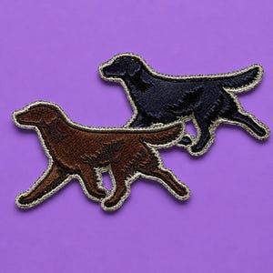 Puede incluir: Dos parches de tela bordados con forma de perros corriendo. Un parche es de color marrón oscuro con un borde claro, y el otro es negro con un borde claro. Ambos parches presentan costuras detalladas que representan la forma del perro.