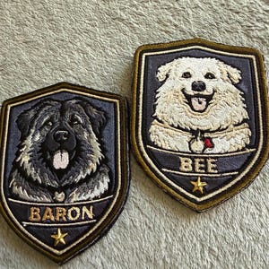 May include: Two embroidered patches featuring dog portraits. One patch shows a black dog with the name "BARON" and a gold star. The other patch depicts a white dog with the name "BEE" and a gold star. Both patches have a shield shape with a gold border.