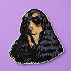 Puede incluir: Un parche bordado que muestra un perro spaniel de color negro y fuego. El perro tiene pelaje largo y oscuro con marcas de color fuego en el hocico, las cejas y el pecho. El parche tiene un borde blanco cosido y sigue el contorno de la cabeza.