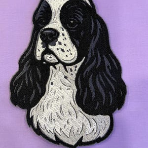 May include: Embroidered patch featuring a black and white Cocker Spaniel dog head. The patch has detailed stitching, showcasing the dog's fur, eyes, and nose. The background is a solid lavender color, providing a contrasting backdrop for the dog's image.