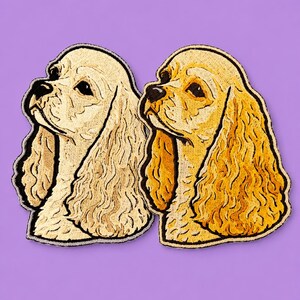 May include: Two embroidered Cocker Spaniel dog patches. One is a light beige, the other a golden yellow. Both have black outlines and are set against a purple background. The patches depict the dog's profile.