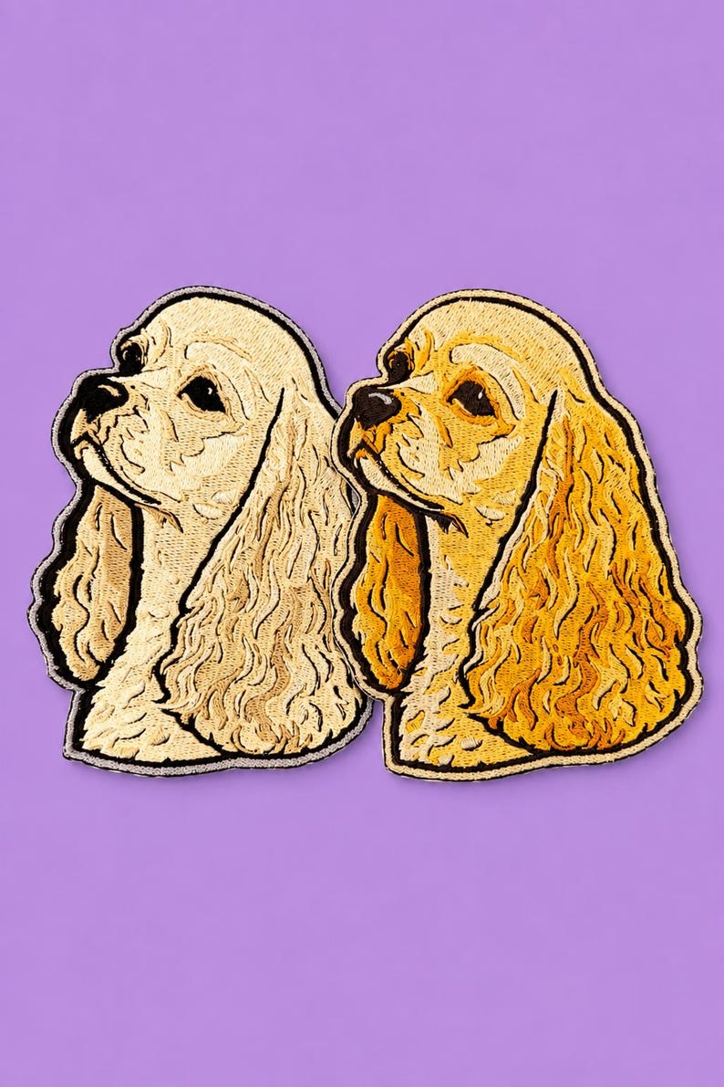 May include: Two embroidered Cocker Spaniel dog patches. One is a light beige, the other a golden yellow. Both have black outlines and are set against a purple background. The patches depict the dog's profile.