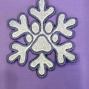 May include: Embroidered snowflake patch with white thread and a light purple outline, set against a solid lavender background. The snowflake design features six symmetrical arms with rounded tips and a detailed centre.