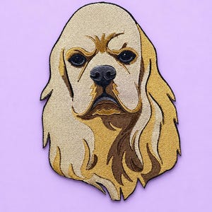 May include: An embroidered patch featuring the head of a golden-colored spaniel dog with long, flowing fur, dark eyes, and a black nose. The design uses shades of tan, cream, and dark brown thread to create depth and detail in the canine portrait.