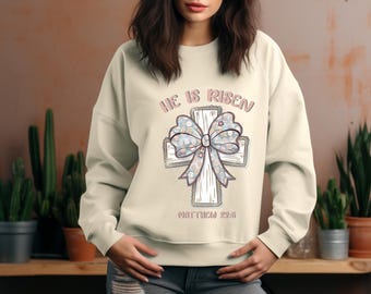 May include: A cream-colored sweatshirt with a floral patterned cross and the text "He is Risen" in pink lettering. The text "Matthew 28:6" is printed below the cross.