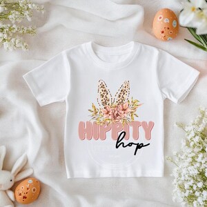 Op de afbeelding: Wit T-shirt met een roze en bruin konijnenorenontwerp en de tekst "Hippity Hop" in roze letters.