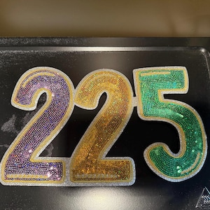 May include: The number 225 is displayed in large, colorful, glittery numbers. The number 2 is purple and gold, the second 2 is gold and green, and the 5 is green and gold.