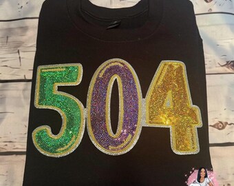 May include: Black sweatshirt with a large sequined number "504" in green, purple, and gold. The sequins are arranged in a pattern that resembles Mardi Gras beads.