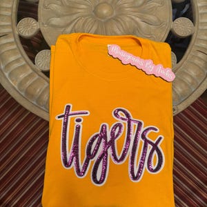 May include: An orange t-shirt with the word "tigers" in a sequined, cursive font. The text is outlined in black and filled with pink sequins. A pink tag that says "Monograms By Ambs" is attached to the shirt.