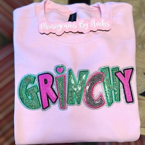 May include: A light pink sweatshirt with the word "GRINCHY" in a sequined design. The letters are outlined in silver glitter, with green and pink sequins. A small pink heart is above the letter "I". A tag that says "Monograms By Ambs" is on the collar.
