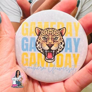 May include: A white round button with the words "Gameday Gameday Gameday Southern University" in yellow and blue lettering. A cartoon jaguar with a fierce expression is in the center of the button.