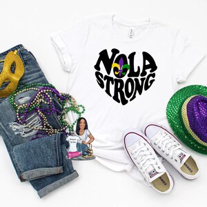 May include: White t-shirt with a black heart-shaped graphic that says "NOLA STRONG" with a fleur-de-lis in the center. The shirt is laying on a white surface with a pair of white sneakers, a pair of jeans, and a Mardi Gras hat.