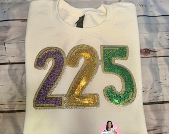 May include: White sweatshirt with the number 225 in purple, gold, and green sequins. The sequins are arranged in a Mardi Gras color scheme.