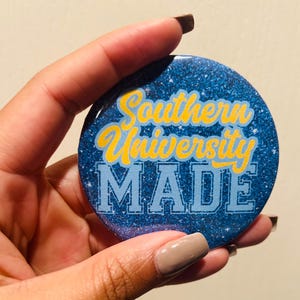 Op de afbeelding: Een ronde, glinsterende blauwe button met de tekst "Southern University MADE" in geel en lichtblauw. De button wordt in een hand gehouden.