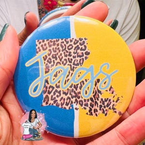 May include: A round, blue and yellow mirror with a leopard print outline of the state of Louisiana and the word "Jaguars" in blue script.