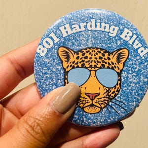 May include: A blue and white button with a leopard wearing sunglasses. The button says "801 Harding Blvd."