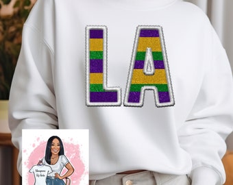 May include: White sweatshirt with a large, purple, green, and gold "LA" graphic on the front. The graphic is outlined in white and has a glittery texture.