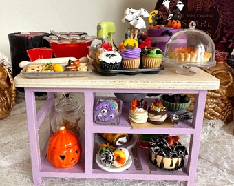 Op de afbeelding: Een miniatuur Halloween-keukeneiland, lavendelkleurig geschilderd, gevuld met kleine bakwaren en decoraties. Cupcakes, koekjes en een taart worden tentoongesteld, samen met een pompoen-lantaarn en een miniatuurmixer. Een doos met mini-kaarsen is zichtbaar op de achtergrond.