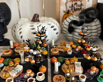 Op de afbeelding: Een Halloween-thema miniatuur voedseldisplay met kleine cakes, taarten en sandwiches op zwarte serveerplanken. Miniatuur pompoenen, kaarsen en decoratieve pompoenen omringen het eten. Een ingelijste foto met een zwarte kat en de woorden "Trick or Treat" staat op de achtergrond.
