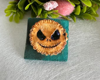 Op de afbeelding: Een kleine, ronde, bruine en zwarte koek met een Jack Skellington gezicht erop. De koek staat op een kleine, vierkante, groene houten basis.