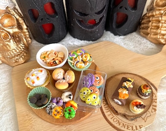 Op de afbeelding: Een houten snijplank met een verscheidenheid aan miniatuur Halloween-thema voedsel, waaronder cupcakes, koekjes, donuts en een rolcake. De plank is versierd met een kleine houten cirkel met het woord "Autionnelie" erop gegraveerd. Er zijn ook drie zwarte kaarsen met rode was en gesneden gezichten op de plank.